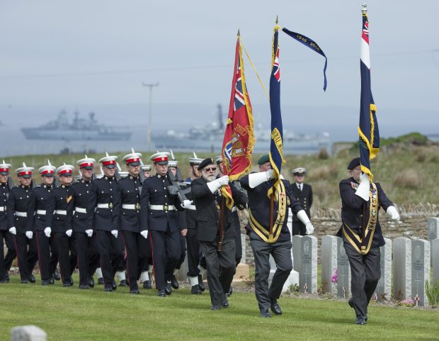 National Commemoration of the Centenary of the Battle of Jutland - Lyness Royal Naval Cemetery on the Island of Hoy. Standard Bearers and Royal Marines march onto the cemetery with HMS Kent and FGS Schleswig-Holstein in the distance.