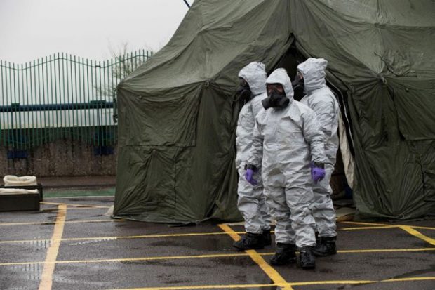 Three individuals are wearing grey personal protective equipment body suits and black face masks. There is a large green tent in the background. The individuals are standing on tarmac.