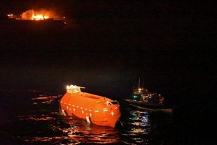 The crew of HMS Argyll, in their Rigid Hull Inflatable Boat, nudge the lifeboat of Grande America towards HMS Argyll, as the cargo ship burns in the background.