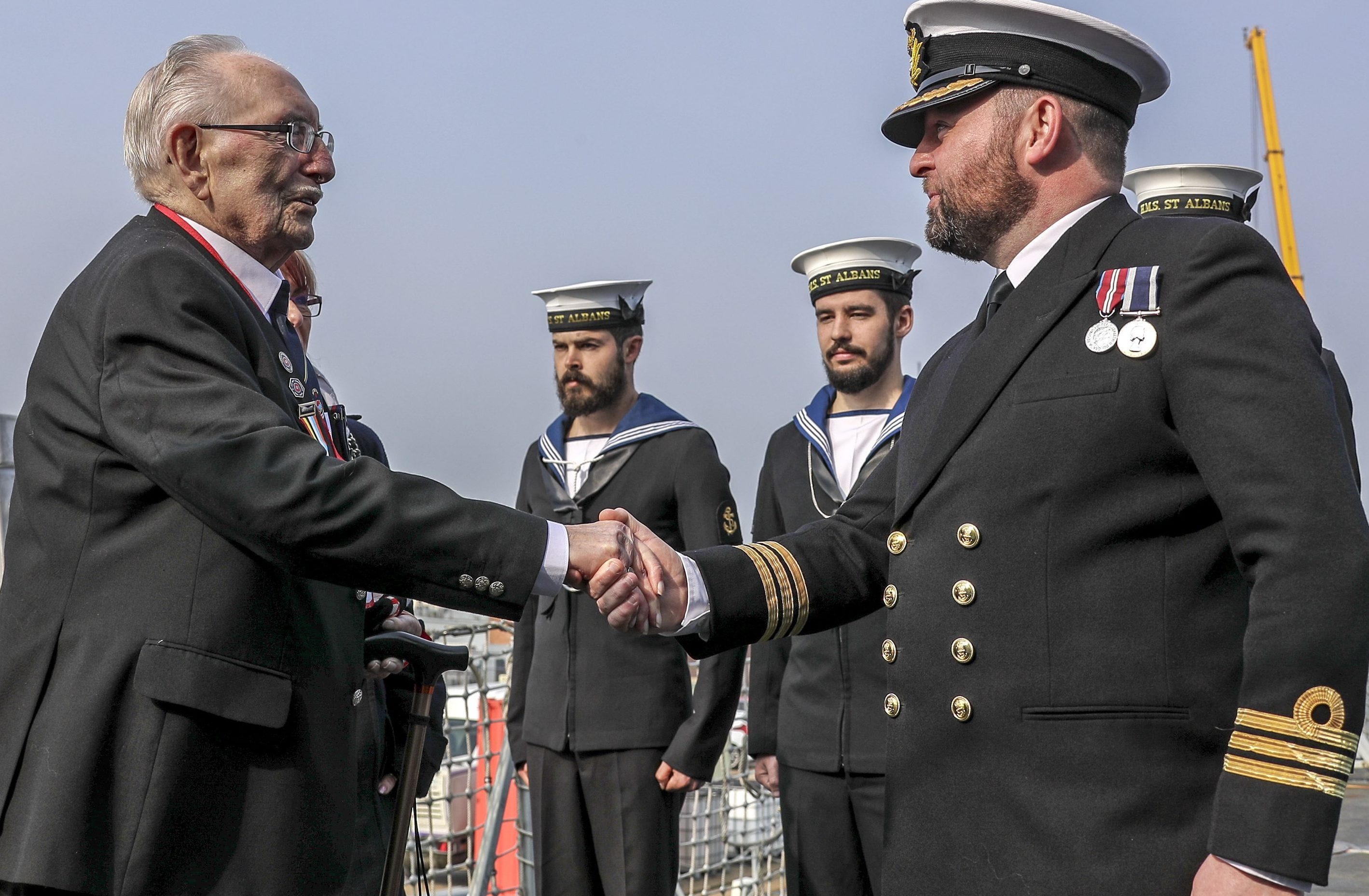 D-Day veteran, Leonard Williams, shakes hands with the Commanding Officer of HMS Saint Albans on board the ship, with two navy sailors standing to attention in the background.