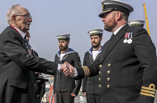 D-Day veteran, Leonard Williams, shakes hands with the Commanding Officer of HMS Saint Albans on board the ship, with two navy sailors standing to attention in the background.