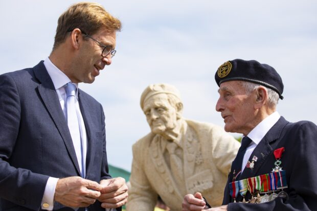 Defence Minister Tobias Ellwood talks to a Normandy veteran at the D-Day Garden. They are in front of a statue of another veteran that is part of the garden.