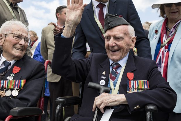 A veteran wearing a military beret and medals smiles and waves from his wheelchair 