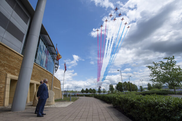 The Prince of Wales watches a formation of Red Arrows flying over the GCHQ building, leaving a red white and blue trail behind them.