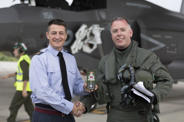 Station Commander, Group Captain Ian Townsend shakes hands with Wing Commander Scott Williams and presents the squadron patch to him in front of an F-35B jet