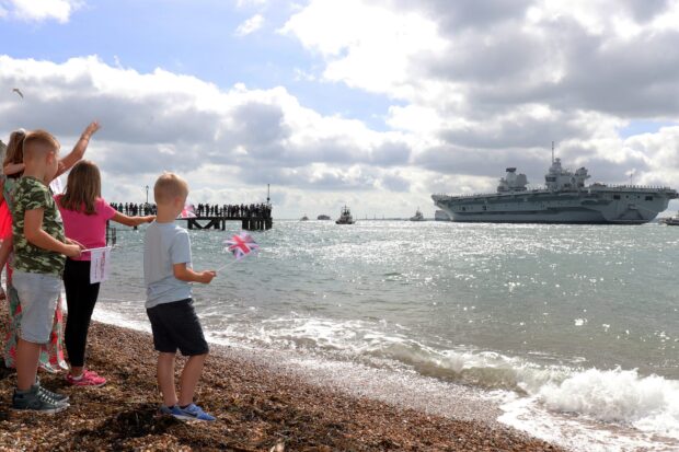 Four children wave off HMS Queen Elizabeth from a beach, while the aircraft carrier sails past in the background